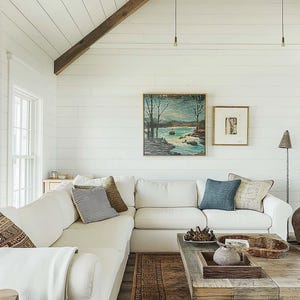 Puede incluir: Un sofá seccional blanco con almohadas y una manta se encuentra frente a una pared blanca de tablones de madera con una pintura de paisaje enmarcada y una pintura abstracta enmarcada más pequeña. Una mesa de café de madera con una cesta tejida y un jarrón de cerámica se encuentra frente al sofá. Una alfombra de patrón marrón está en el suelo.