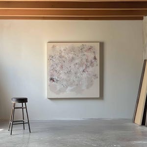 May include: A white canvas painting with abstract brushstrokes in shades of pink, gray, and white hangs on a white wall in a studio setting. A black metal stool sits in the foreground.