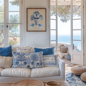 May include: A white sofa with blue and white patterned cushions in a living room with a view of the sea. The sofa is in front of a large window with white French doors. There is a wooden coffee table with a woven tray and a brown ceramic bowl.