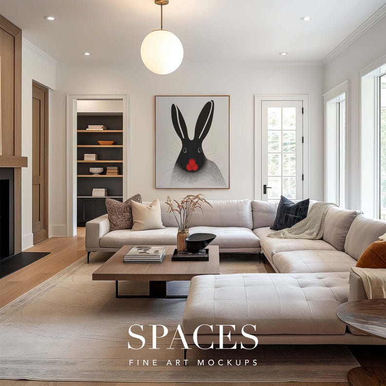 May include: A white sectional sofa with a black and white rabbit art print hanging above it. The sofa is in a living room with a large window and a wooden door. There is a coffee table in front of the sofa with a book and a vase of flowers on it.