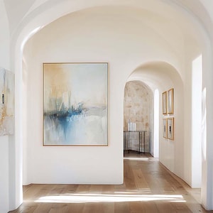 Puede incluir: Una pintura abstracta en un marco dorado cuelga de una pared blanca en un pasillo. La obra de arte presenta tonos de azul, beige y blanco. El pasillo tiene puertas arqueadas y un suelo de madera. Se ven obras de arte enmarcadas adicionales.