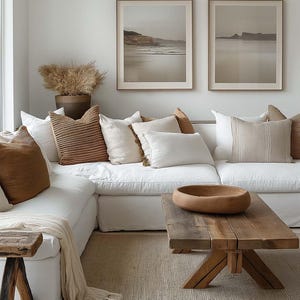 May include: A white sectional sofa with brown throw pillows and a blanket, a wooden coffee table with a brown bowl, and a rug on the floor. Two framed landscape photos hang on the wall.