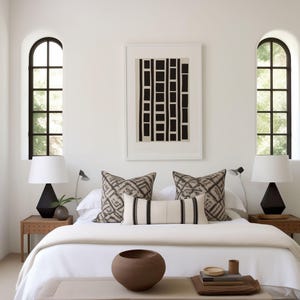 Puede incluir: Una cama blanca con ropa de cama blanca y almohadas con estampado en blanco y negro. La cama está en una habitación con paredes blancas y dos ventanas. Hay un banco de madera delante de la cama con un cuenco marrón y una pila de libros encima.
