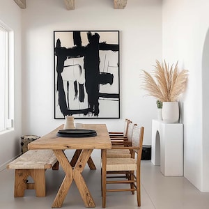 Peut inclure: Salle à manger avec une grande table en bois et un banc assorti, ainsi que des chaises à assise tressée. Une peinture abstraite en noir et blanc est accrochée au mur. Des herbes de la pampa dans un vase blanc.