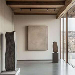 May include: An interior shot of a modern art gallery with sculptures and a large painting. The room features a polished concrete floor, neutral-toned walls, and a wooden ceiling with track lighting. Large windows offer a view of the landscape.