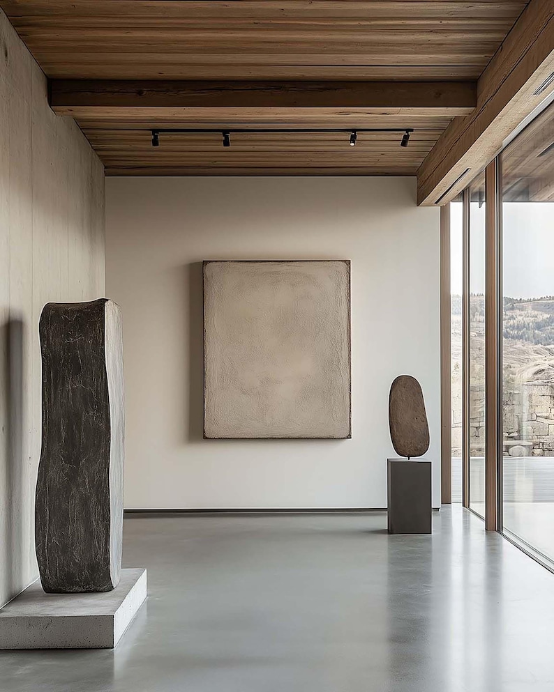 May include: An interior shot of a modern art gallery with sculptures and a large painting. The room features a polished concrete floor, neutral-toned walls, and a wooden ceiling with track lighting. Large windows offer a view of the landscape.