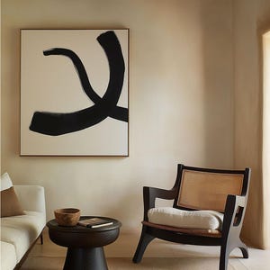 May include: A black and white abstract painting hangs on a beige wall above a dark wood side table with a bowl and a book. A black wicker chair with a white cushion sits in the corner of the room.
