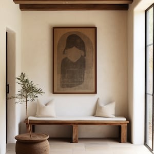 Puede incluir: Escena interior de tonos neutros con un banco de madera, dos cojines color crema y una obra de arte enmarcada. Un pequeño árbol en una maceta tejida está a la izquierda. La habitación tiene un diseño minimalista con luz natural.