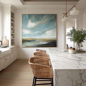 Puede incluir: Interior de cocina con una pintura de paisaje que representa un cielo con nubes. Tres taburetes de bar de ratán tejido con patas de metal negro están colocados en una encimera de mármol. La habitación tiene gabinetes blancos, pisos de madera y una lámpara decorativa.