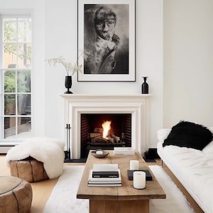 Könnte beinhalten: Ein weißes Wohnzimmer mit einem Kamin, einem weißen Sofa, einem weißen Schaffellteppich und einem Holz-Couchtisch mit Büchern und Kerzen. Ein Schwarzweißporträt hängt über dem Kamin.