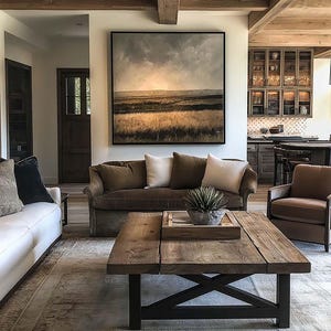 Pode incluir: Uma sala de estar com uma pintura de paisagem acima de um sofá marrom e almofadas brancas. Uma mesa de centro rústica de madeira com base preta repousa sobre um tapete estampado. Assentos adicionais incluem um sofá branco e uma poltrona marrom.