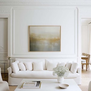 May include: A light and spacious living room featuring white walls, a white sofa, and a square coffee table. An abstract painting in a gold frame hangs above the sofa. A vase of white flowers sits on the table. The room has a minimalist design.