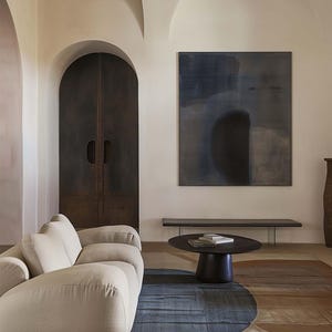 May include: A light beige, curved sofa with a dark wood coffee table and a black and white abstract painting on the wall. The room has a vaulted ceiling and a large arched doorway.