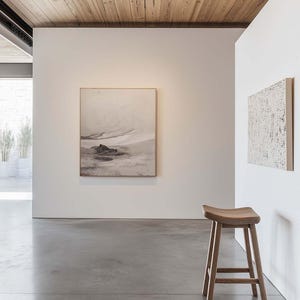Può includere: Un interno moderno con soffitto in legno chiaro e pavimento in cemento. Un'opera d'arte paesaggistica incorniciata è appesa a una parete bianca, accanto a un'opera d'arte strutturata. Uno sgabello in legno con seduta curva aggiunge calore naturale allo spazio.