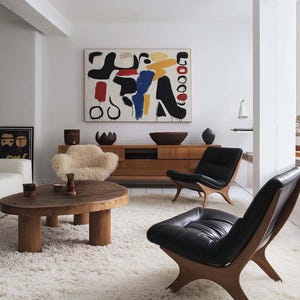 Puede incluir: Un interior de sala de estar con una alfombra blanca, una mesa de café de madera, dos sillas de cuero negro con marcos de madera y una pared blanca con una pintura abstracta en negro, rojo, amarillo, azul y blanco.
