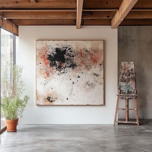 Peut inclure: Une peinture abstraite blanche avec des éclaboussures de peinture noire, rouge et brune est accrochée à un mur blanc dans une pièce avec un sol en béton et des poutres en bois. Un chevalet se trouve dans le coin de la pièce.