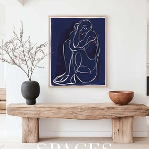 Puede incluir: Un banco de madera con una impresi&oacute;n enmarcada en azul oscuro de un dibujo lineal de una persona sobre un fondo blanco. La impresi&oacute;n est&aacute; colgada en una pared blanca. Un peque&ntilde;o jarr&oacute;n con una rama seca est&aacute; en el banco. Un cuenco de madera est&aacute; en el banco a la derecha de la impresi&oacute;n. El texto "SPACES FINE ART MOCKUPS" est&aacute; en la parte inferior de la imagen.