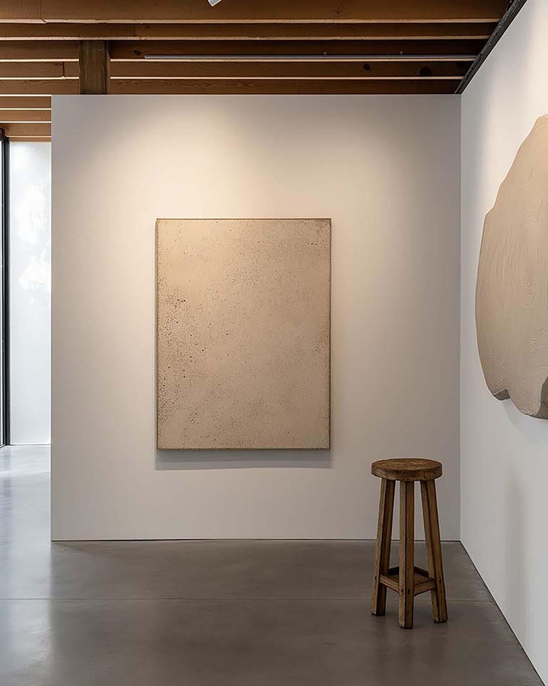 May include: An art gallery interior featuring a neutral-toned, textured rectangular artwork on a white wall. A small, round wooden stool with three legs sits on the polished concrete floor. The space is well-lit, with exposed wooden beams on the ceiling.