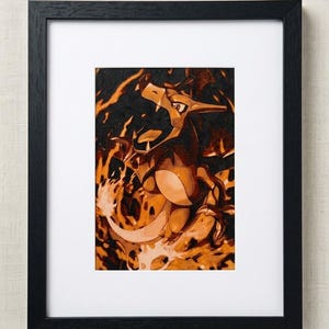 May include: A framed art print featuring a stylized orange and brown dragon-like creature with an open mouth, surrounded by swirling orange and yellow flames. The artwork is encased in a simple black frame with a white mat border.