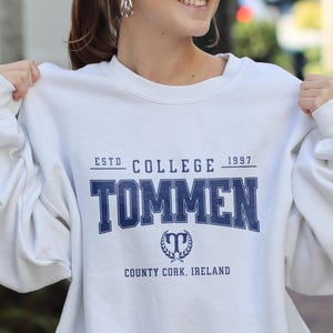 May include: White crewneck sweatshirt with the text "TOMMEN" in navy blue block letters. The sweatshirt also features the text "ESTD COLLEGE 1997" and "COUNTY CORK, IRELAND" in navy blue. A crest is centered below the word "TOMMEN".