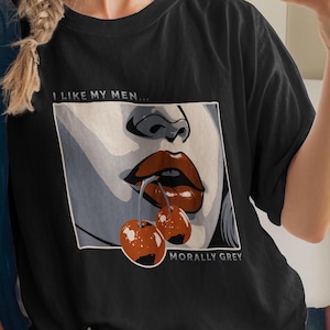 May include: Black t-shirt with a graphic of a mouth and cherries, with the text "I LIKE MY MEN... MORALLY GREY". The shirt is worn by a person wearing a blue cap and sunglasses.