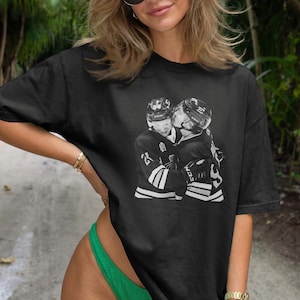 May include: Black t-shirt featuring a grayscale image of two hockey players embracing. The players are wearing hockey helmets and jerseys. The t-shirt is worn by a person wearing sunglasses and a green swimsuit.