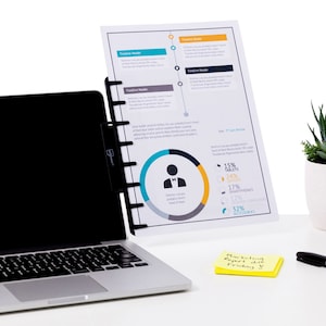 May include: A laptop with a black screen and a document holder attached. A printed document with charts and graphs is held in the holder. A yellow sticky note with text is on the desk, alongside a pen.