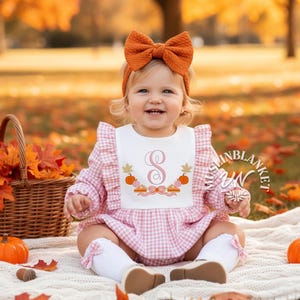 Puede incluir: Un bebé con un pelele de cuadros rosas y blancos con un babero blanco bordado con una inicial y calabazas. El bebé también lleva una diadema con lazo naranja y está sentado sobre una manta con calabazas y hojas de otoño.