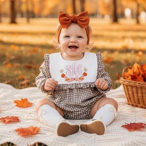 Może przedstawiać: Niemowlę w brązowym rompie w kratę, białym śliniaku z monogramem i opasce z kokardą w kolorze rdzy. Śliniak ma haftowany wzór z dyniami i gwiazdami. Scena rozgrywa się na zewnątrz z jesiennymi liśćmi i dyniami.