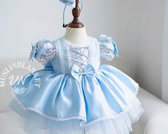 Puede incluir: Vestido de bebé azul claro con diadema a juego. El vestido presenta un corpiño de satén con detalle de cordones, mangas con lentejuelas y una falda amplia sobre una enagua de tul. Un gran lazo adorna la cintura. La diadema tiene un lazo.