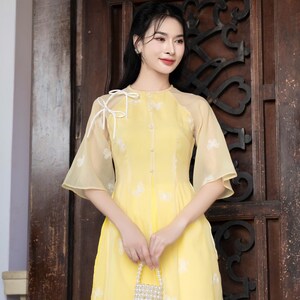 Puede incluir: Una mujer con un vestido amarillo con bordados florales blancos y un lazo blanco en el hombro. Lleva un pequeño bolso de cuentas blancas.