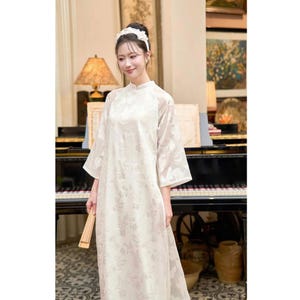 Puede incluir: Un vestido color crema con estampado floral, cuello mandarín y mangas tres cuartos. El vestido se lleva con una diadema a juego. Se sostiene un abanico plegado en la mano. El fondo incluye un piano y obras de arte.