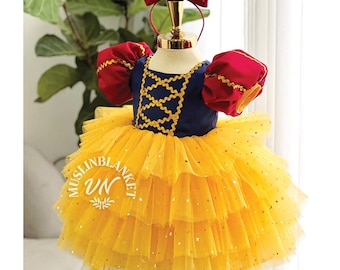May include: A yellow tulle dress with gold glitter, a blue bodice with gold trim, and red puff sleeves. The dress is worn by a mannequin with a red bow headband. The dress is labeled "Muslinblanket VN".