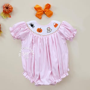 May include: A pink baby girl's smocked dress with short sleeves and a ruffled hem. The dress has an embroidered collar with a ghost, pumpkin, spider, and candy corn design.