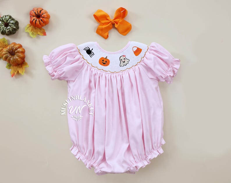 May include: A pink baby girl's smocked dress with short sleeves and a ruffled hem. The dress has an embroidered collar with a ghost, pumpkin, spider, and candy corn design.