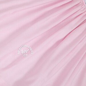 May include: A close-up of a pale pink fabric with a white embroidered logo that reads "Muslin Blanket UN".