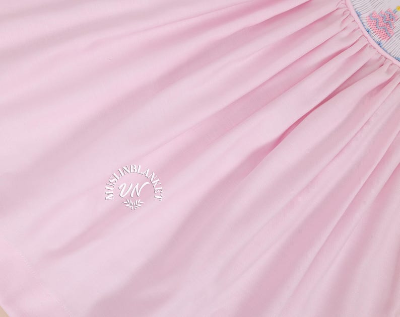 May include: A close-up of a pale pink fabric with a white embroidered logo that reads "Muslin Blanket UN".