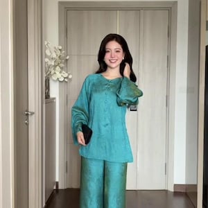 May include: A teal two-piece outfit. The top has long sleeves and a button-down front. The matching trousers are wide-legged. The fabric has a subtle sheen. The outfit is worn in a well-lit hallway.