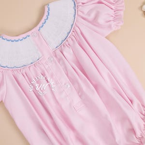 May include: A pink baby smocked dress with a white and blue smocked collar and short puffed sleeves. The dress has a button closure in the back and a small embroidered logo on the back.