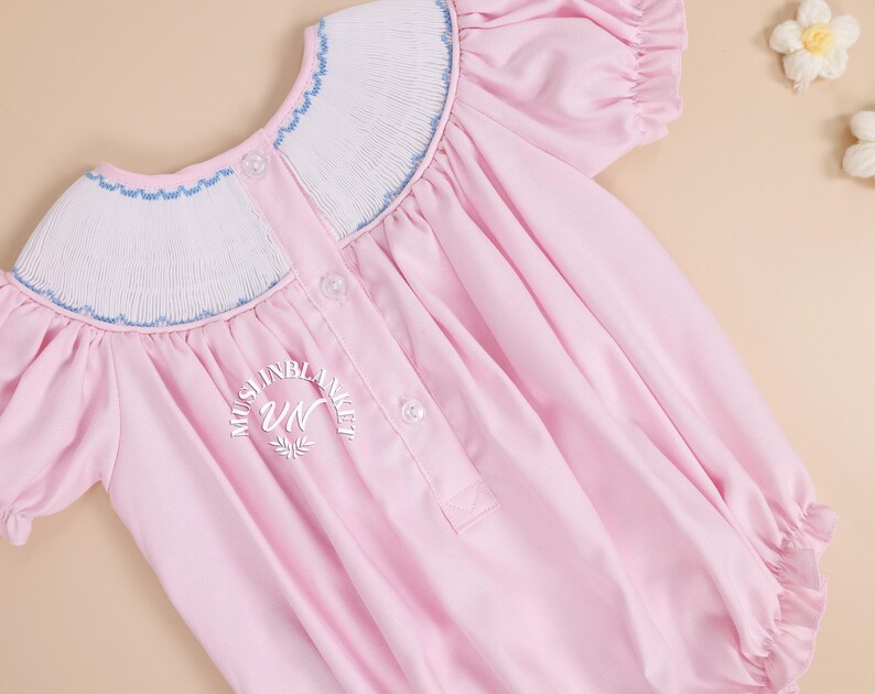 May include: A pink baby smocked dress with a white and blue smocked collar and short puffed sleeves. The dress has a button closure in the back and a small embroidered logo on the back.