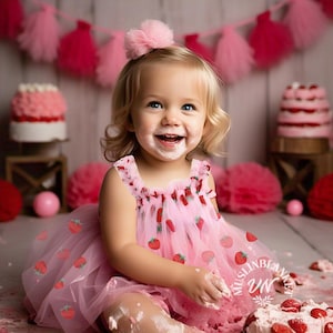 May include: A young girl wearing a pink tulle dress with strawberry print sits in front of a cake, covered in frosting. She is smiling and has frosting on her face.