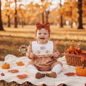 Può includere: Un bambino con una fascia con fiocco color ruggine e un pagliaccetto a quadretti abbinato con un bavaglino bianco. Il bavaglino è ricamato con un monogramma e disegni di tacchini. La scena comprende zucche, foglie d'autunno e un cestino di vimini.