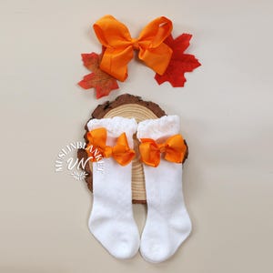 May include: A pair of white baby socks with orange bows. The socks are on a wooden surface with a fall-themed leaf.