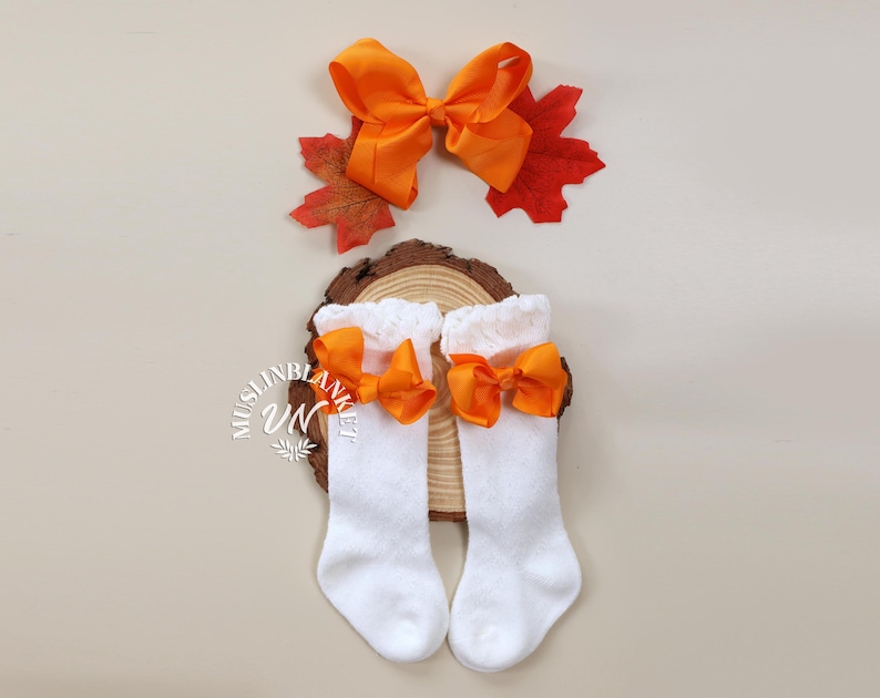 May include: A pair of white baby socks with orange bows. The socks are on a wooden surface with a fall-themed leaf.