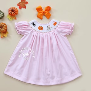 May include: A pink smocked dress with a white embroidered collar featuring Halloween-themed designs, including a pumpkin, ghost, spider, and candy corn. The dress has short puffed sleeves and a gathered skirt.