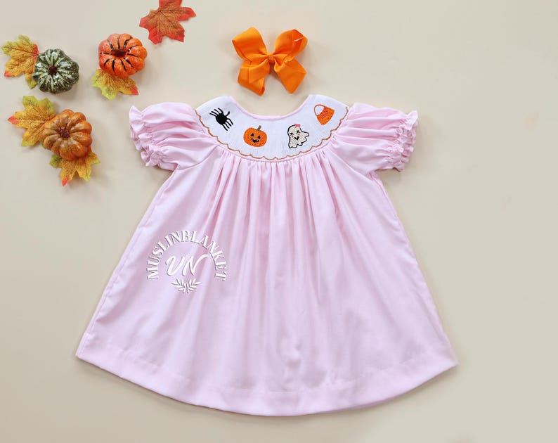 May include: A pink smocked dress with a white embroidered collar featuring Halloween-themed designs, including a pumpkin, ghost, spider, and candy corn. The dress has short puffed sleeves and a gathered skirt.