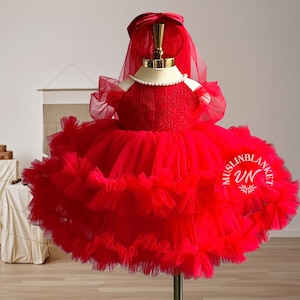 May include: A red tulle dress with a ruffled skirt and a sheer overlay. The dress has a beaded bodice and a bow on the top.