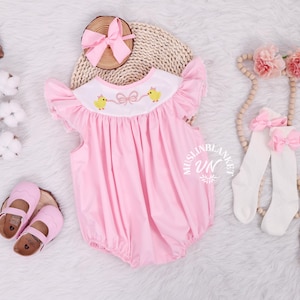Puede incluir: Conjunto rosa para bebé que incluye un pelele con bordado de pollitos amarillos, mangas con volantes y una diadema con lazo rosa a juego. El conjunto se completa con zapatos rosas y calcetines altos blancos con lazos rosas.