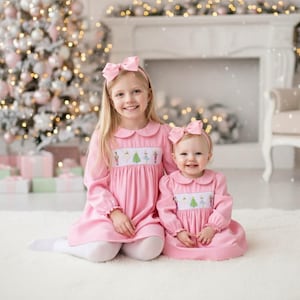Op de afbeelding: Twee jonge meisjes in bijpassende roze jurken met lange mouwen en Peter Pan-kragen, elk met een strik. De jurken hebben geborduurde ontwerpen. Ze zitten voor een kerstboom.