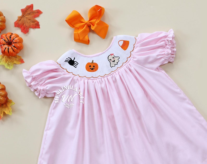 May include: A pink smocked dress with short sleeves and a Halloween-themed embroidered neckline featuring a ghost, pumpkin, spider, and candy corn. The dress is on a light brown background.